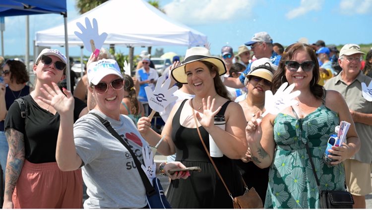 A group of smiling people wave at the camera, holding white hand-shaped signs, enjoying a sunny outdoor event.