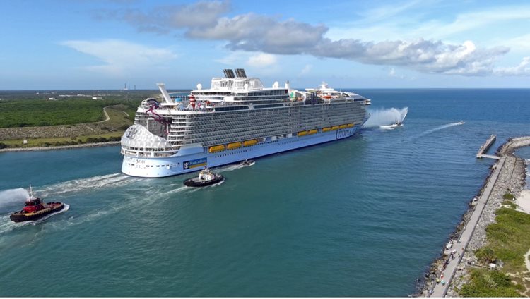 A large cruise ship navigates a waterway, accompanied by tugboats, with a scenic coastline in the background.